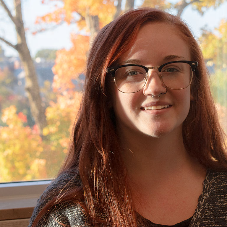 headshot of a woman with red hair and glasses with fall trees in the background